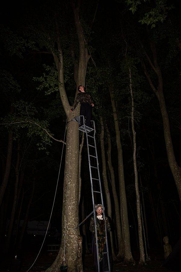 Photography of composer BIlly Bultheel and Alexander Iezzi. It is nighttime in the woods and stands at the top of a long metal ladder that leads to a blind. Alexander Iezzi is on the ground peering through to bars on the ladder.