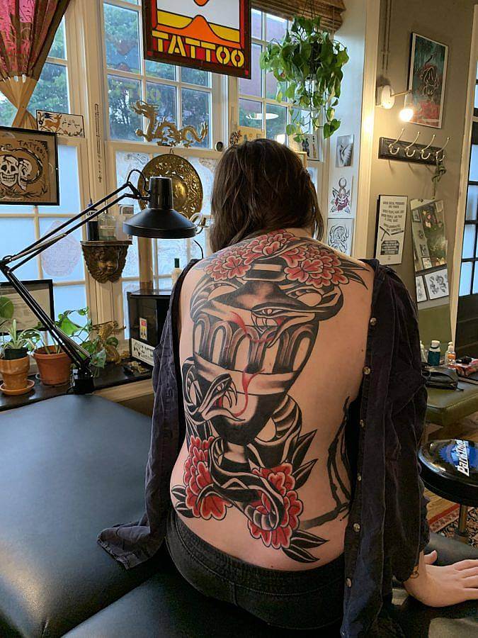 A person sits on a tattoo bed with their back to the camera. On their back is a massive black tattoo of a vase. Two snakes encircle it. Red flowers flanks it at the bottom and top.