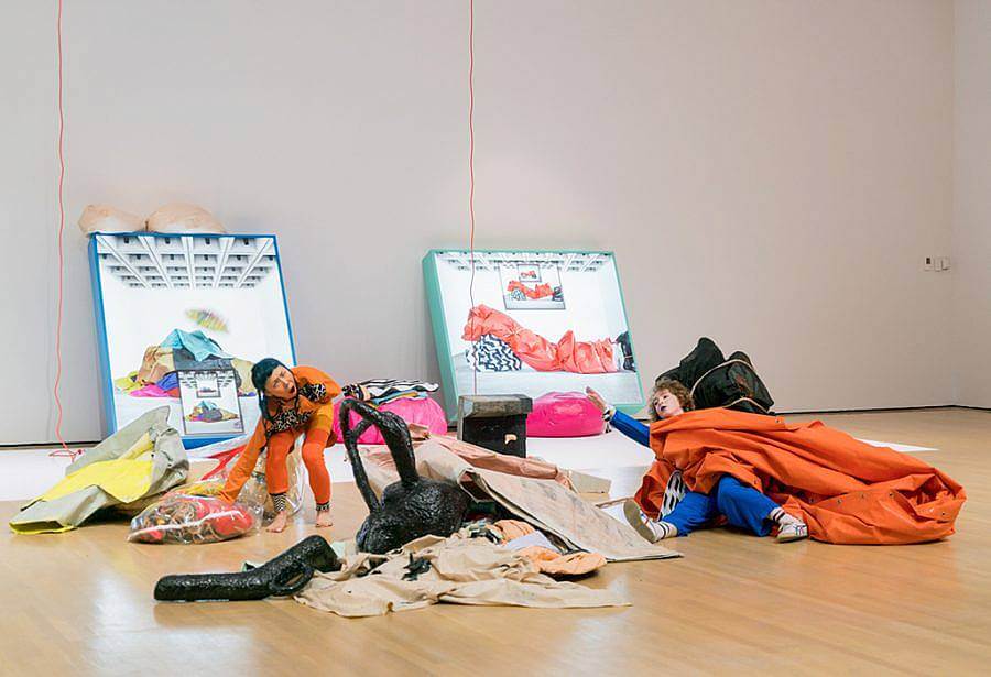 Two performers are depicted. The one of the left grabs a transparent bags with objects in it, the one on the right with are stretched out, lays on the floor enmeshed in objects and fabrics.