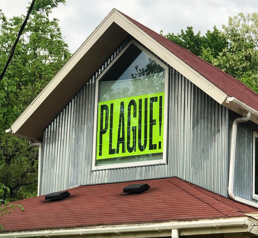 Photo of a two story house. Focused in on a center top window. In the window is a printed sign that says "PLAGUE!" in all caps, it its bolded against a green sheet. The sky is visible behind the house.