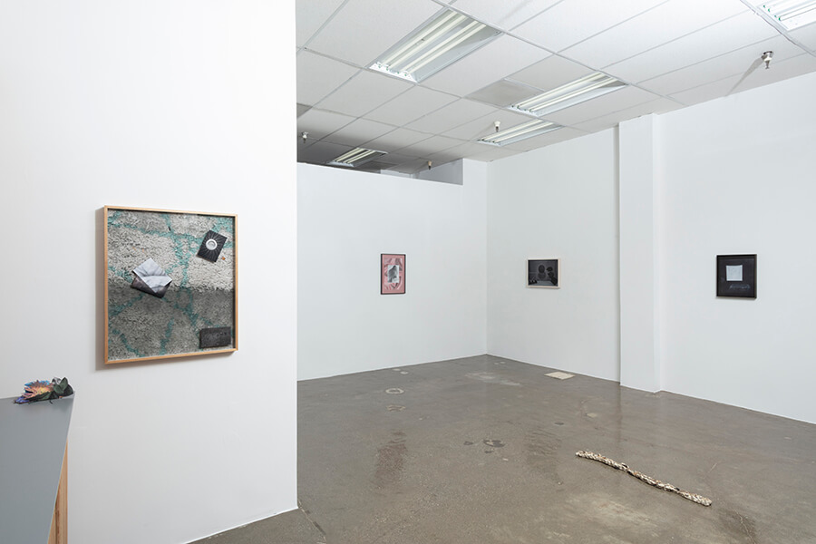 image description: This image is an installation view of After Afterlife at the Fulcrum. The gallery has three walls visible, there. are several framed, rectangular works hung on each wall. There is also a linear sculpture that resmebles a stick that is on the floor. The gallery has white walls, a paneled ceiling, and cement floors.
