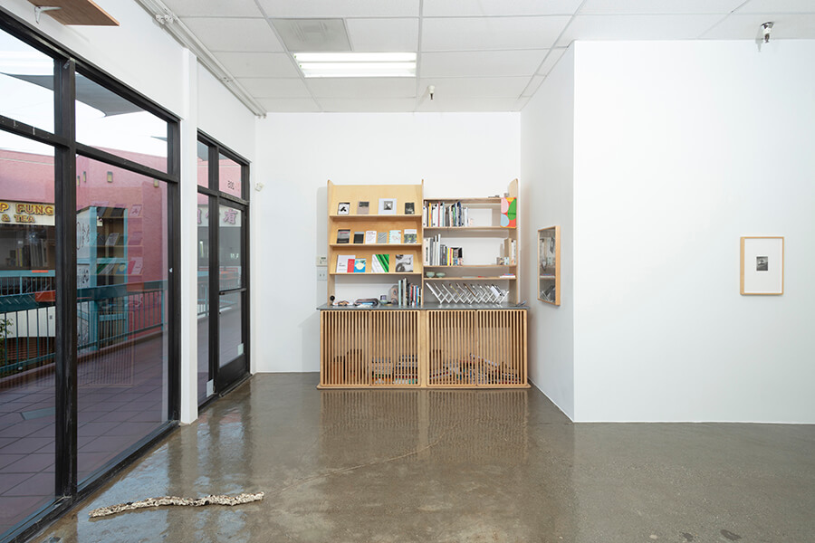 Image description: This is an installation view of the exhibition "After Afterlife" at The Fulcrum Press. In the gallery there is a sculpture on the floor that resembles a stick and two works hanging in rectangular frames that are made from bare wood. The bookshelf is also visible along the back wall. It is made of light wood and features cabinets and slanted shelves to display the front covers of the books.